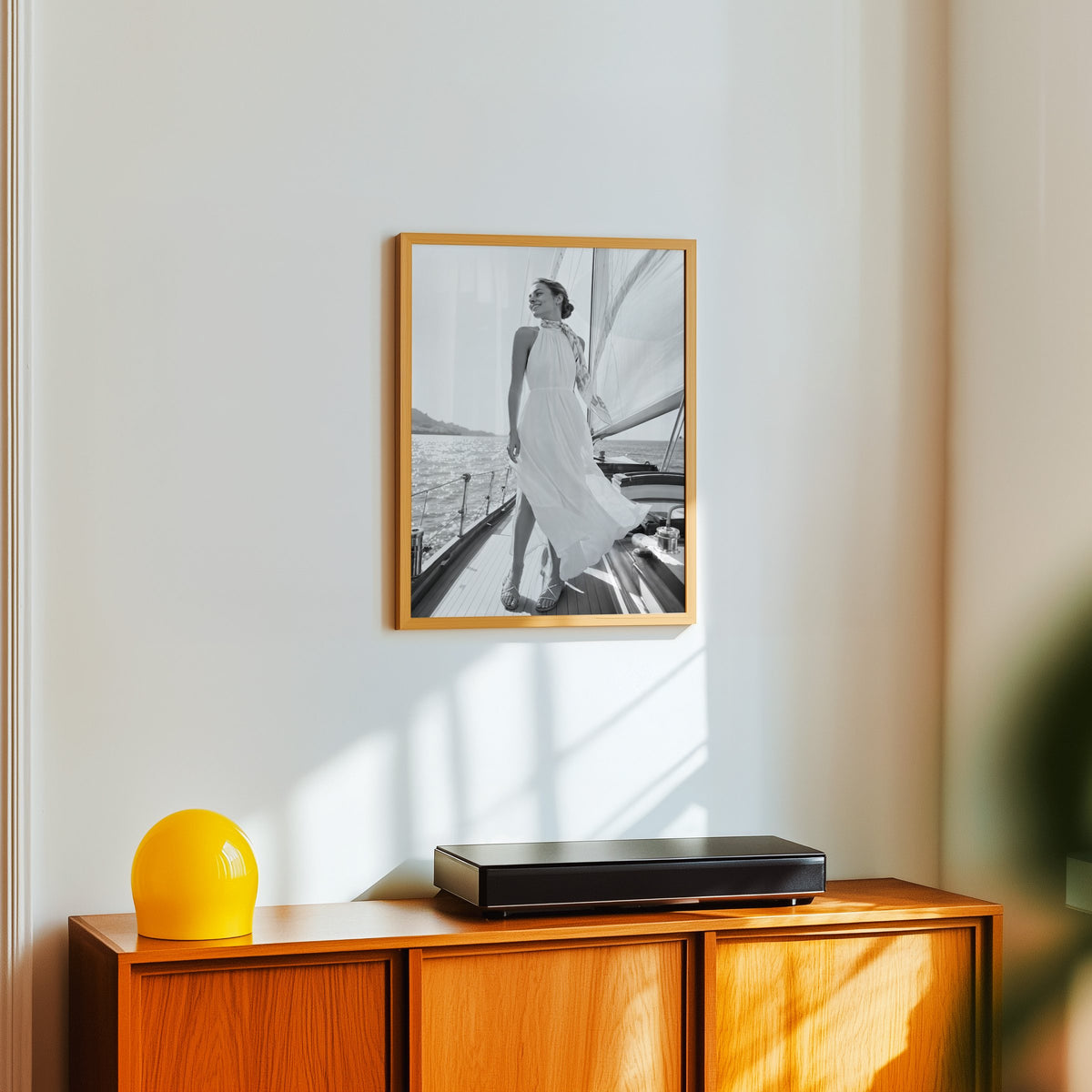 Black and white woman in white dress on sailboat print in an oak frame above a teak sideboard with a yellow glass lamp