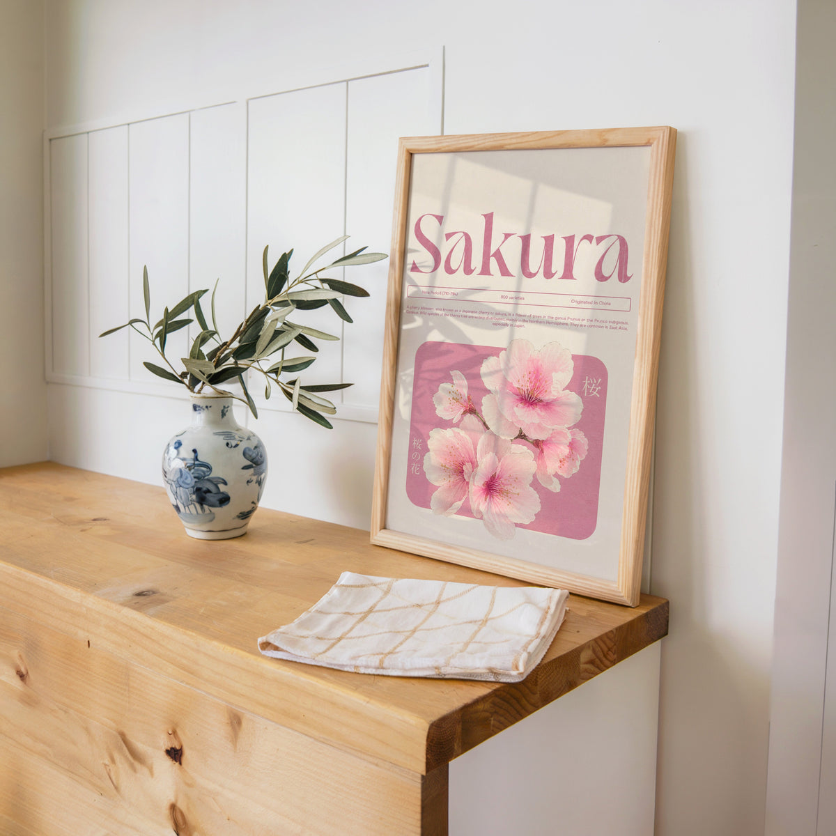 pink sakura blossom typography print in a pine frame on a wooden counter beside a blue and white porcelain vase