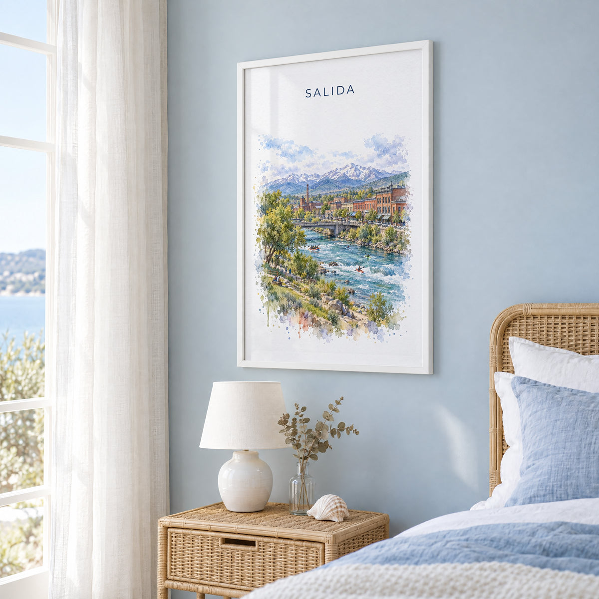 Salida, Colorado watercolor print in a white frame on a pale blue wall beside a rattan bed in a coastal bedroom