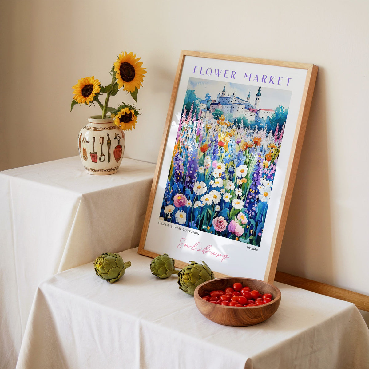 Salzburg flower market print in an oak frame leaning on a linen-draped table beside a vase of sunflowers