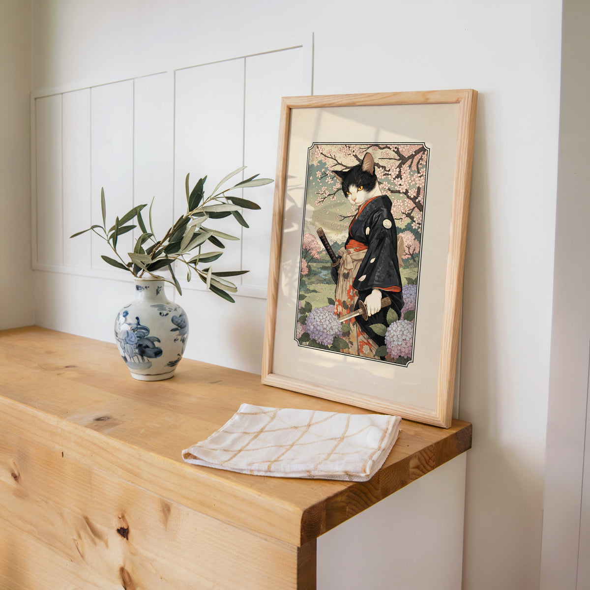 samurai cat with katana print in a pine frame on a wooden counter beside a blue and white porcelain vase