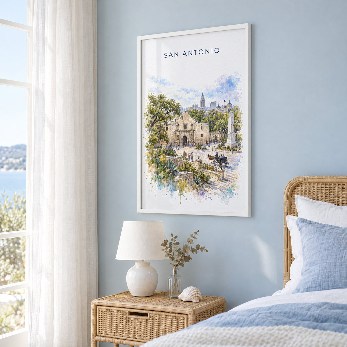 San Antonio watercolor print in a white frame on a pale blue wall beside a rattan bed in a coastal bedroom