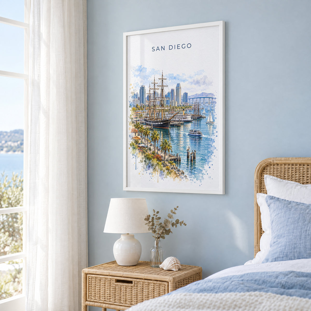 San Diego Bay watercolor print in a white frame on a pale blue wall beside a rattan bed in a coastal bedroom