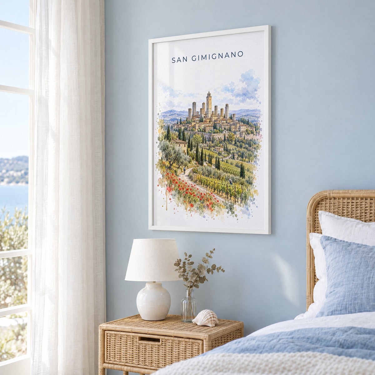 San Gimignano watercolor print in a white frame on a pale blue wall beside a rattan bed in a coastal bedroom