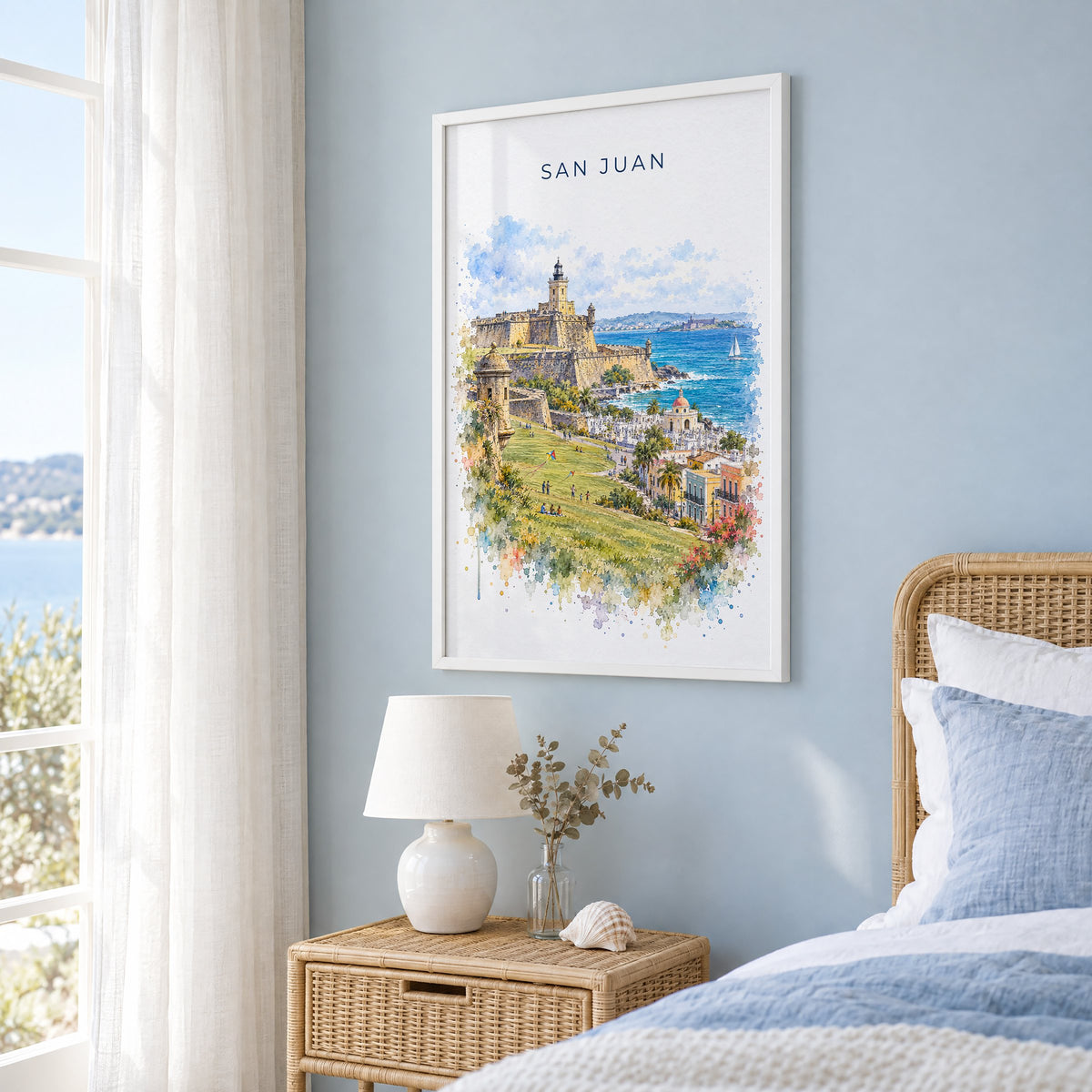 San Juan watercolor print in a white frame on a pale blue wall beside a rattan bed in a coastal bedroom