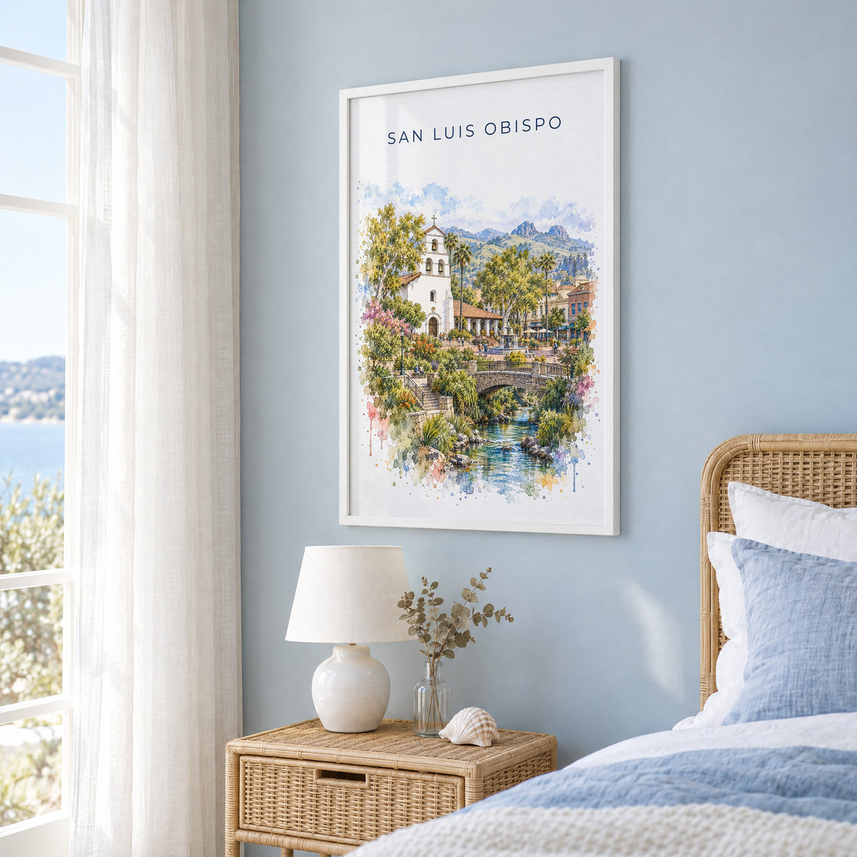 San Luis Obispo watercolor print in a white frame on a pale blue wall beside a rattan bed in a coastal bedroom