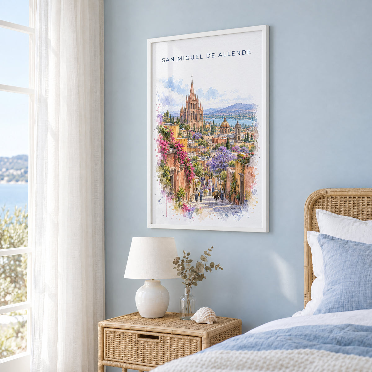 San Miguel de Allende watercolor print in a white frame on a pale blue wall beside a rattan bed in a coastal bedroom