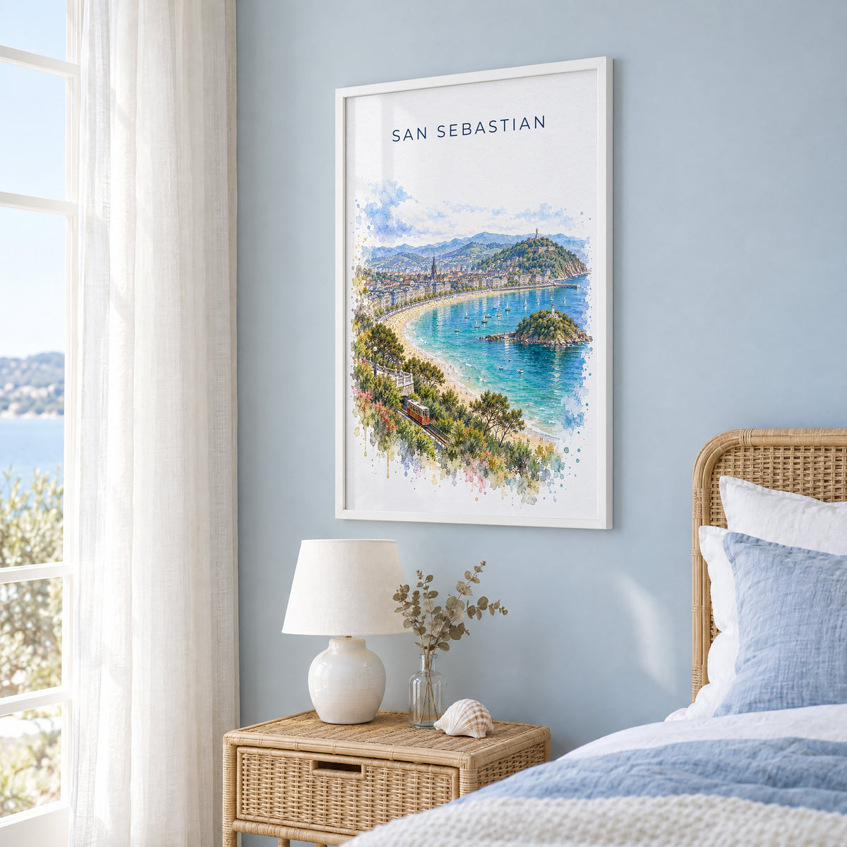San Sebastian watercolor print in a white frame on a pale blue wall beside a rattan bed in a coastal bedroom