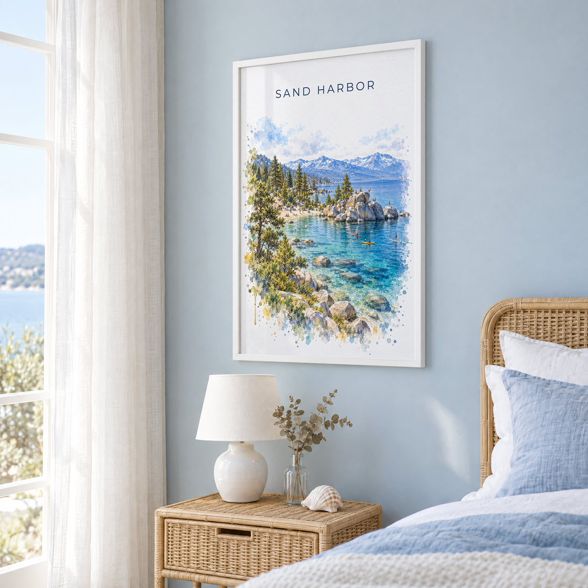 Sand Harbor watercolor print in a white frame on a pale blue wall beside a rattan bed in a coastal bedroom