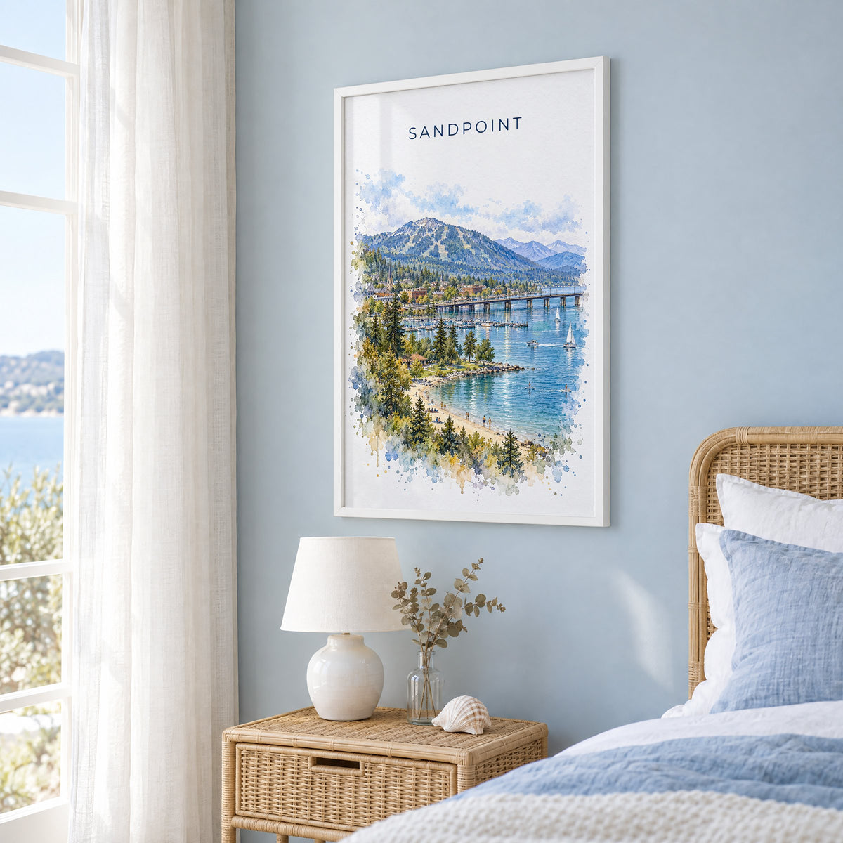 Sandpoint watercolor print in a white frame on a pale blue wall beside a rattan bed in a coastal bedroom