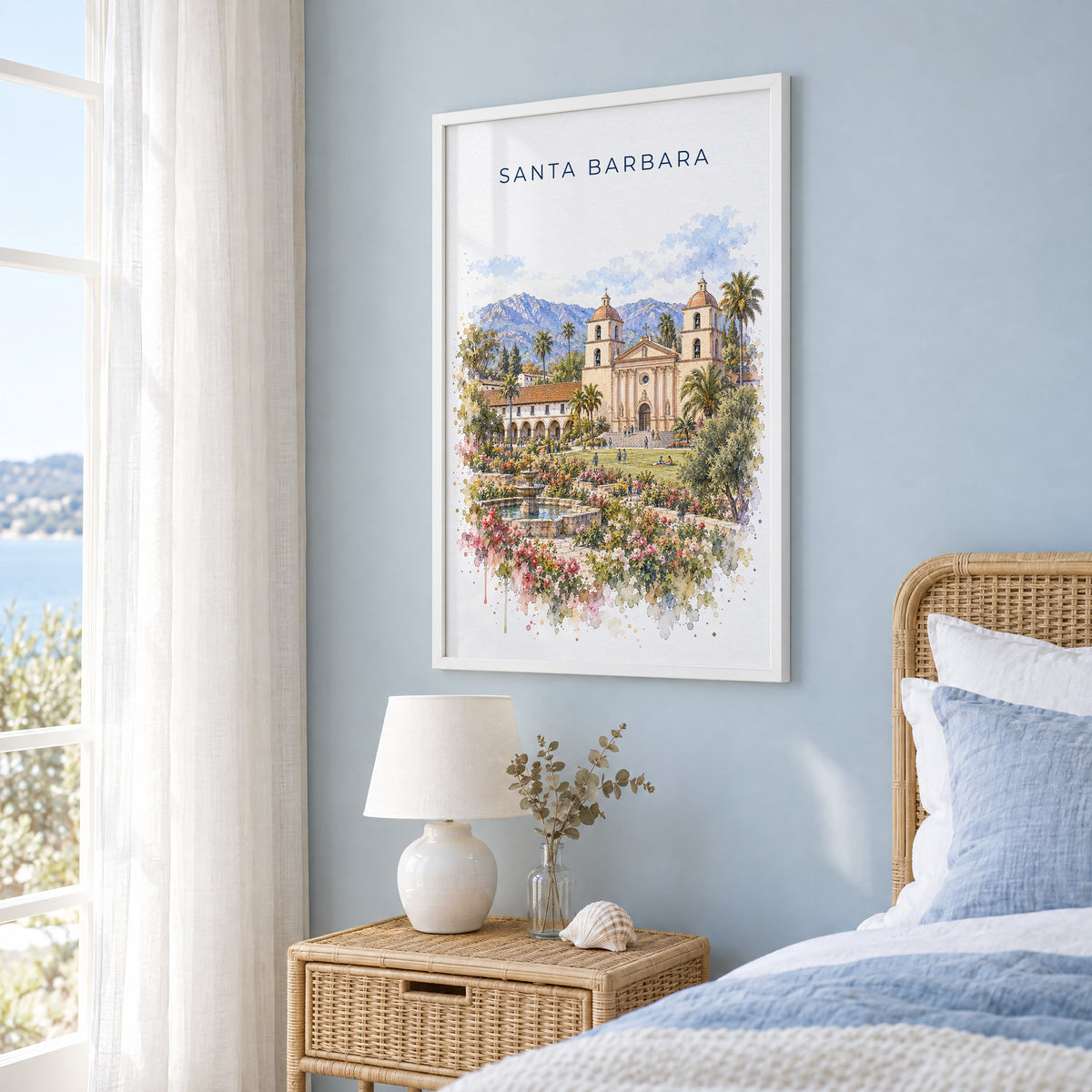 Santa Barbara watercolor print in a white frame on a pale blue wall beside a rattan bed in a coastal bedroom