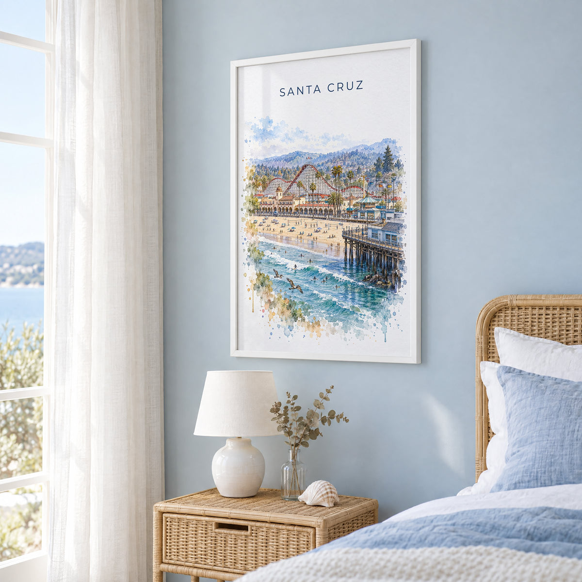 Santa Cruz, California watercolor print in a white frame on a pale blue wall beside a rattan bed in a coastal bedroom