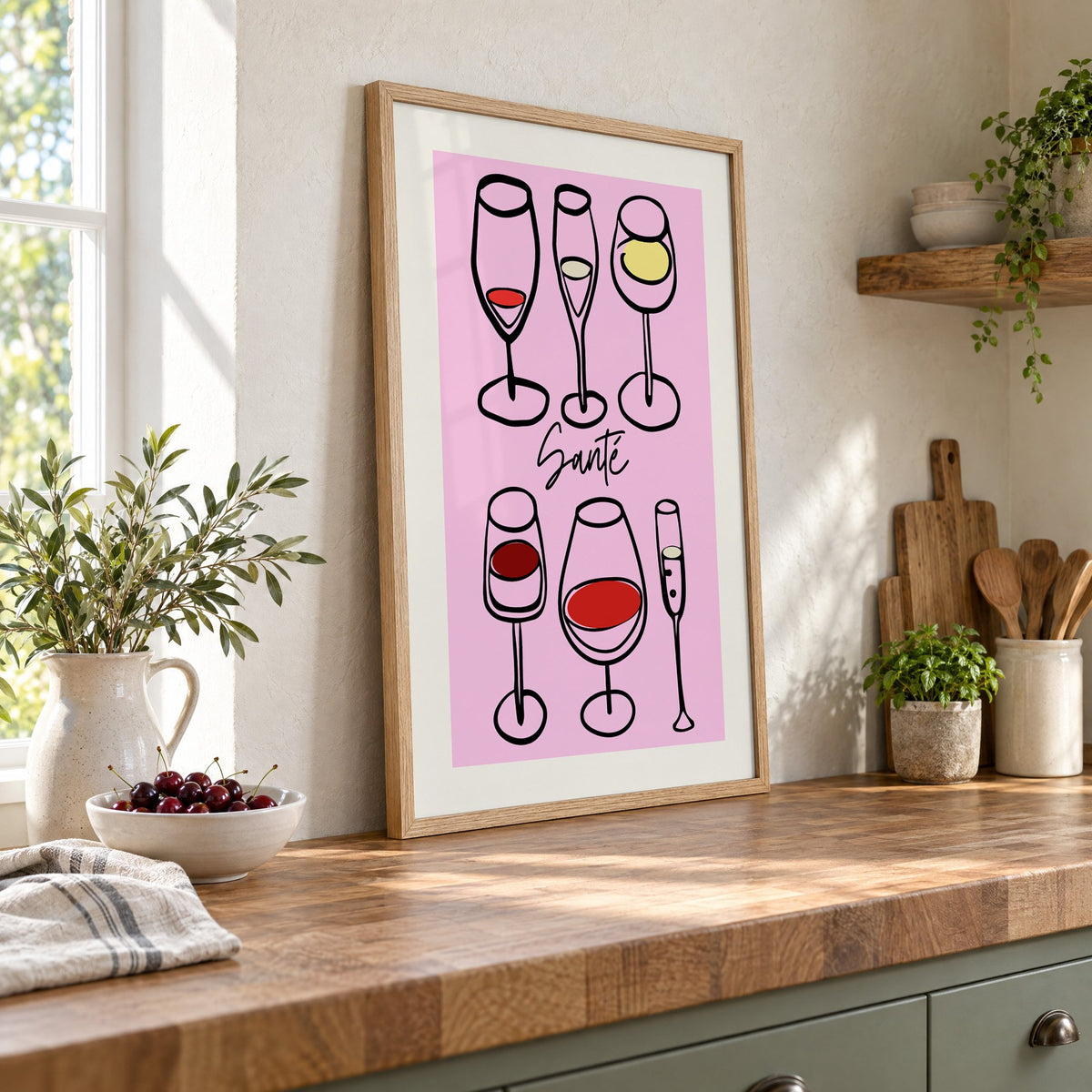 Santé wine glasses line art print in an oak frame leaning on a butcher block island beside a bowl of cherries