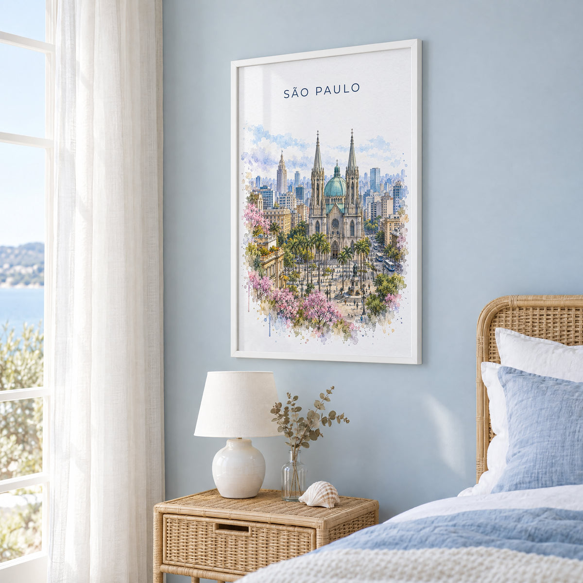 São Paulo watercolor print in a white frame on a pale blue wall beside a rattan bed in a coastal bedroom