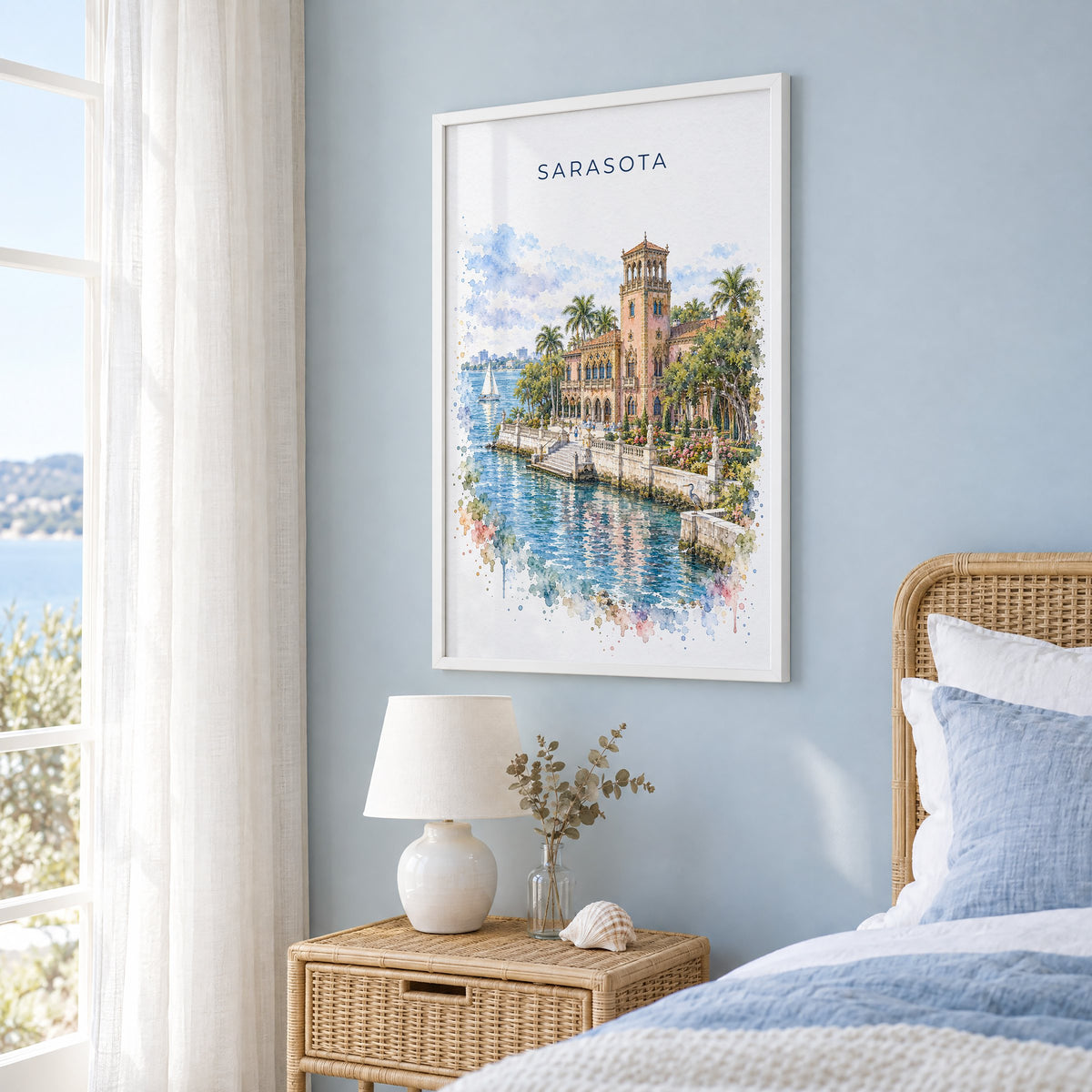 Sarasota watercolor print in a white frame on a pale blue wall beside a rattan bed in a coastal bedroom