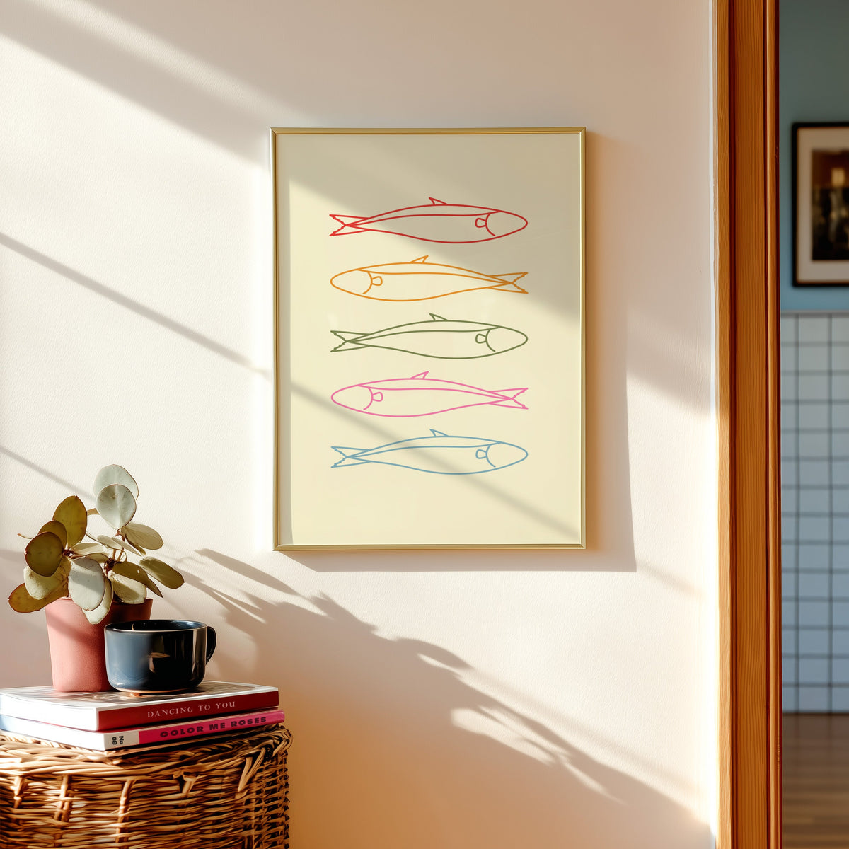 Pastel sardine print shown in a gold frame above a basket of books and a pilea plant