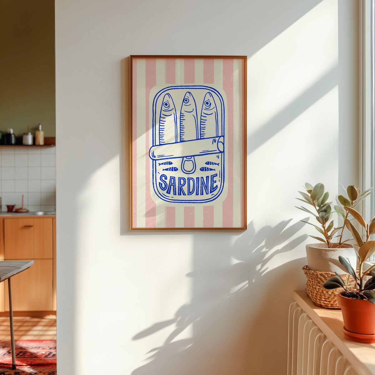 Sardine tin print shown in a gold frame in sunlight with plants and a kitchen beyond