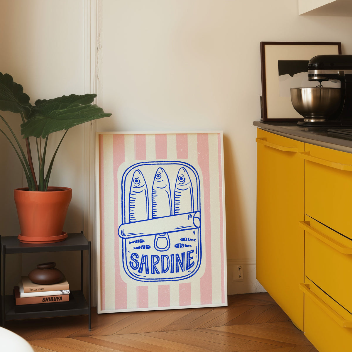 Sardine tin print in a white frame leaning on parquet beside a bright yellow kitchen