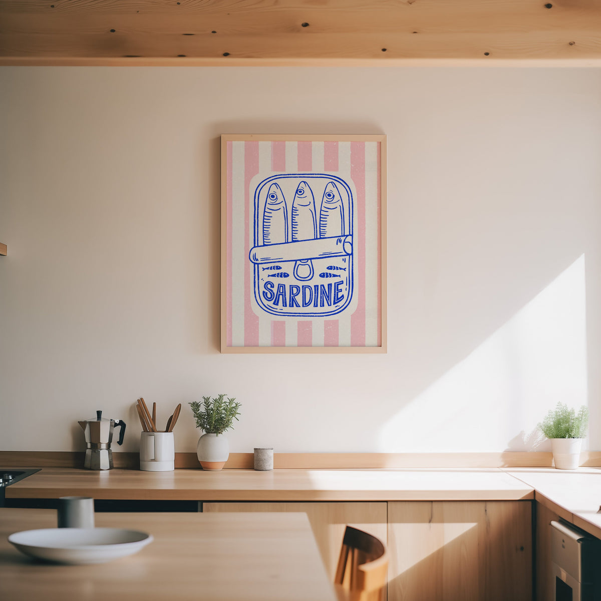 Sardine tin print in an oak frame above a wood counter with a moka pot and herbs