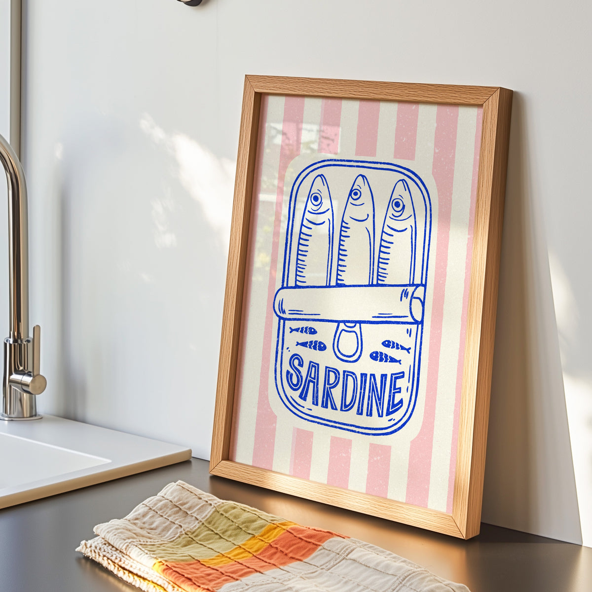 Sardine tin print in an oak frame by the kitchen sink beside a striped tea towel