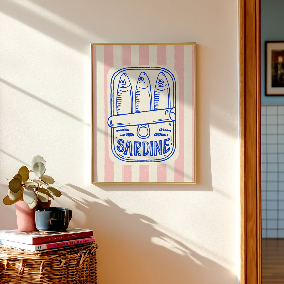 Sardine tin print in a gold frame above a basket of books and a pilea plant