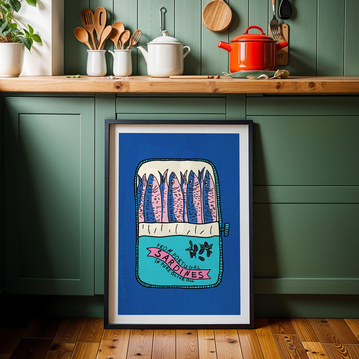 Blue sardine tin print in a black frame against sage green kitchen cabinets