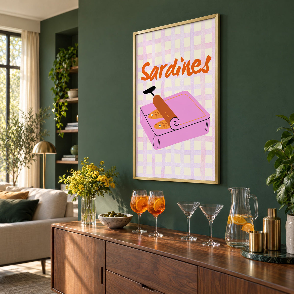 Sardines pink tin on gingham print in a brass frame on a bottle green wall above a sideboard with spritz and martini glasses, seen at an angle