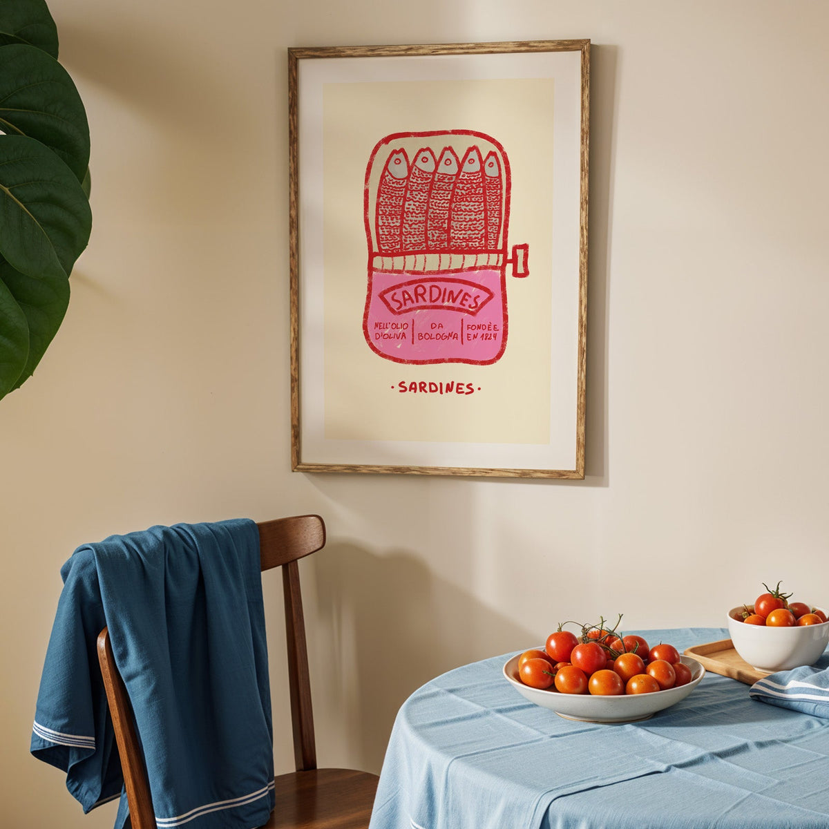 Pink sardine tin print in an oak frame above a blue linen table with ripe tomatoes