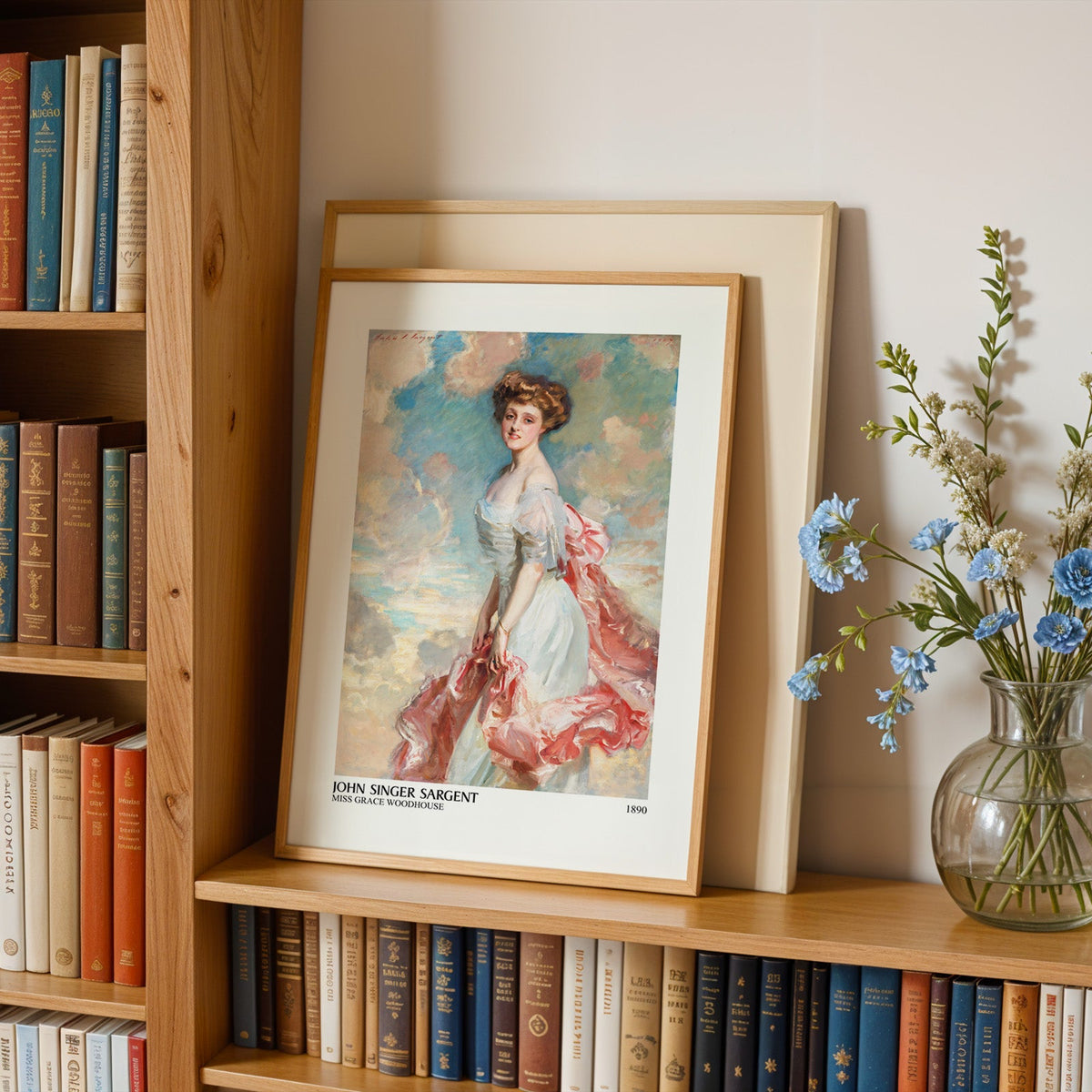 Pink and cream portrait print in an oak frame leaning on a bookshelf beside flowers