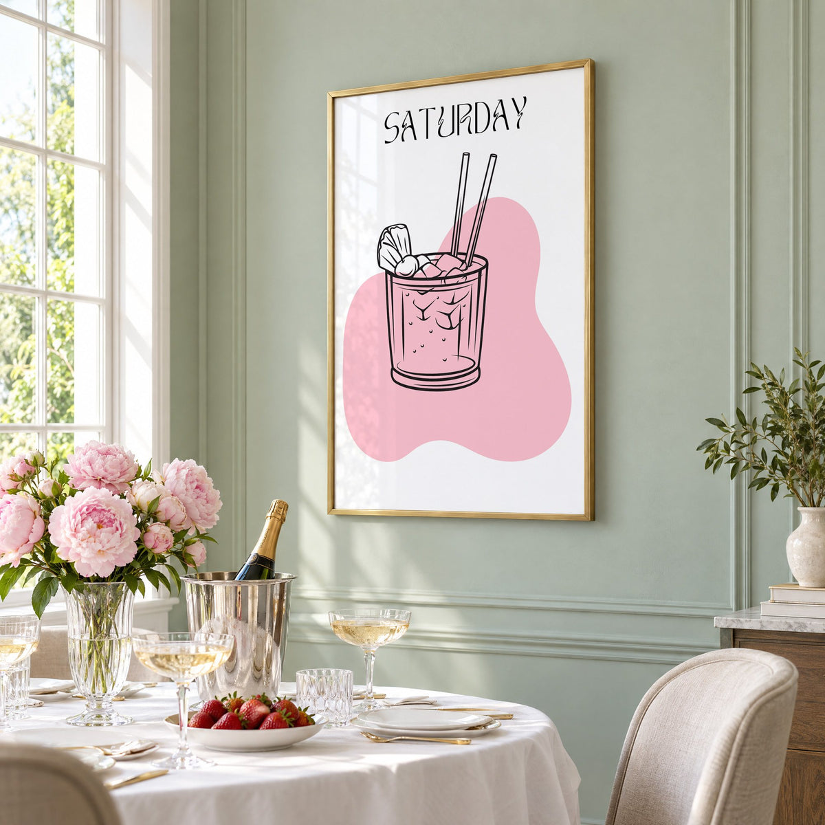 SATURDAY iced cocktail two straws print in a brass frame on a sage wall above a brunch table with champagne and peonies, seen at an angle
