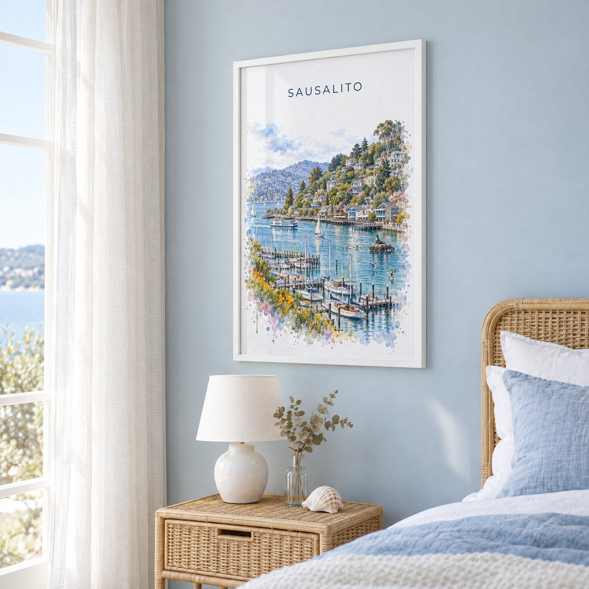 Sausalito watercolor print in a white frame on a pale blue wall beside a rattan bed in a coastal bedroom