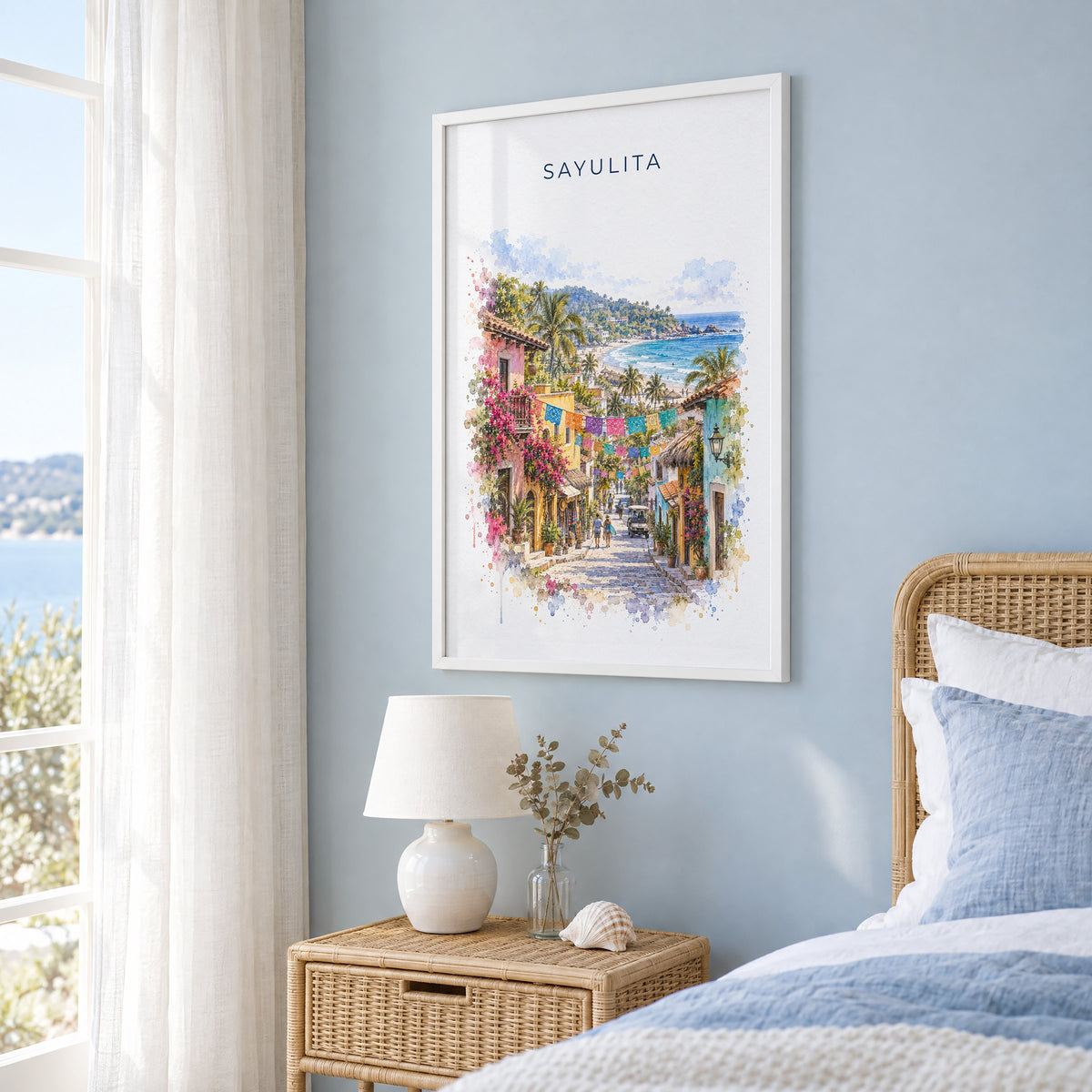 Sayulita watercolor print in a white frame on a pale blue wall beside a rattan bed in a coastal bedroom