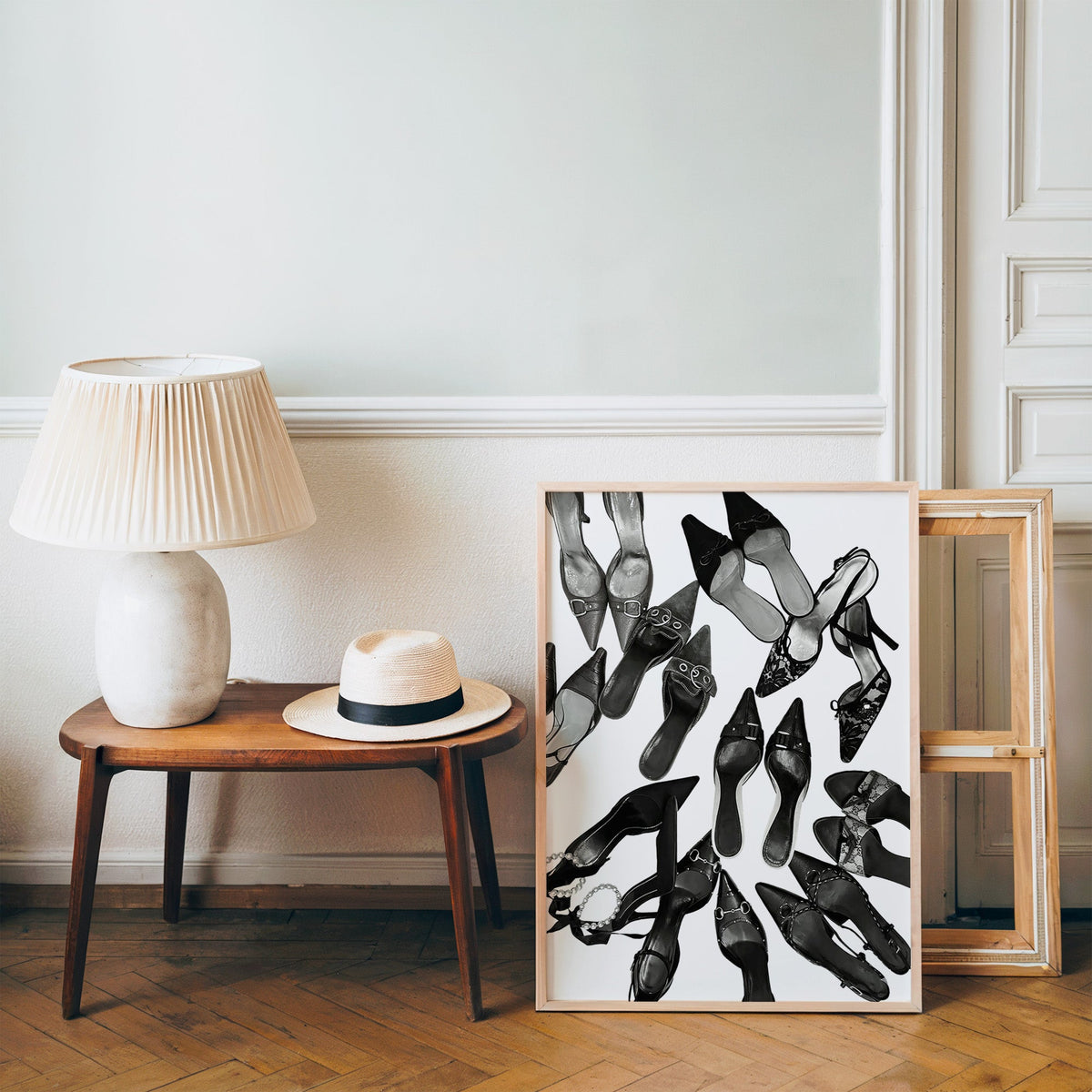 Top-down scattered pointed heels and mules print in a pine frame on a wood floor beside a lamp and straw hat