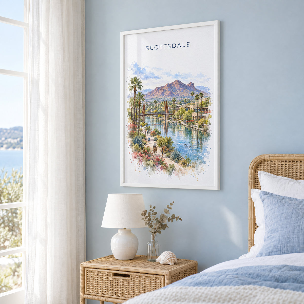 Scottsdale watercolor print in a white frame on a pale blue wall beside a rattan bed in a coastal bedroom