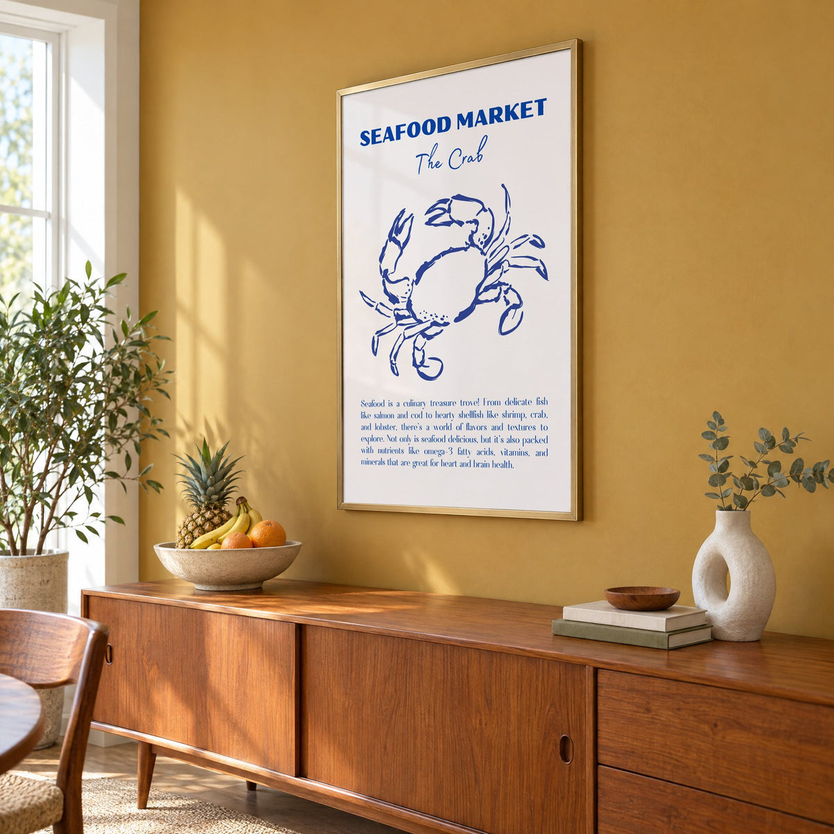 SEAFOOD MARKET The Crab cobalt sketch print in a brass frame on a mustard wall above a teak sideboard with a fruit bowl, seen at an angle