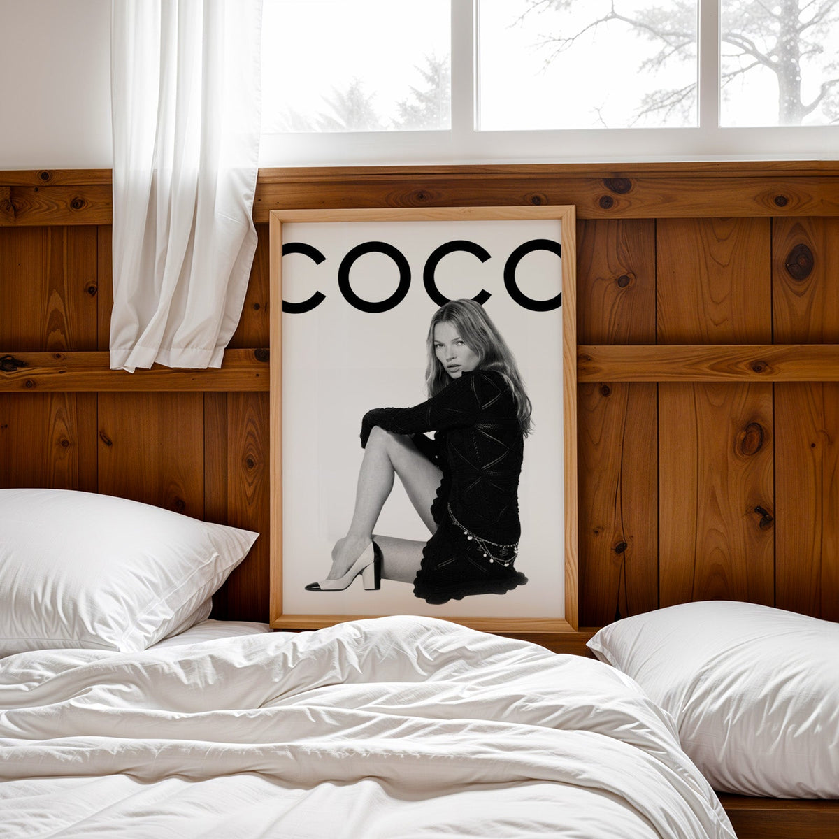 Seated woman in black knit dress under giant letters print in a pine frame leaning on a wood headboard over white bedding