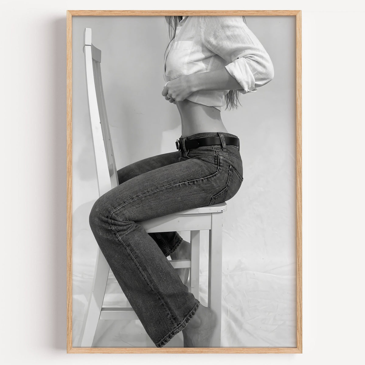 Woman in flared jeans on a white wooden chair print in a light oak frame on a plain white wall