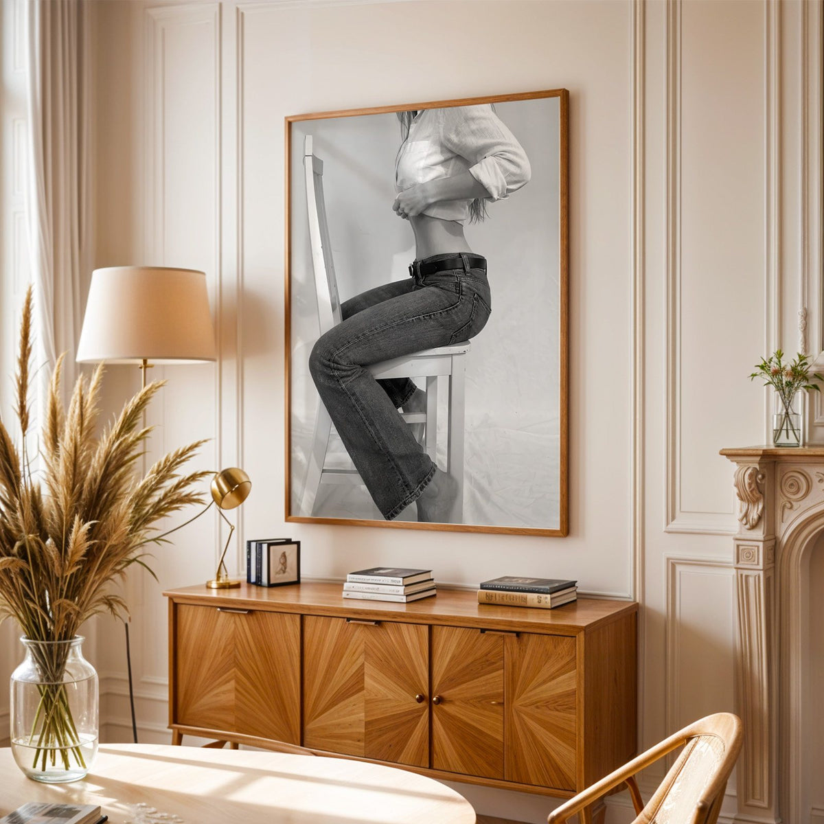 Woman in flared jeans on a white wooden chair print in an oak frame above a sunburst wood sideboard with pampas grass