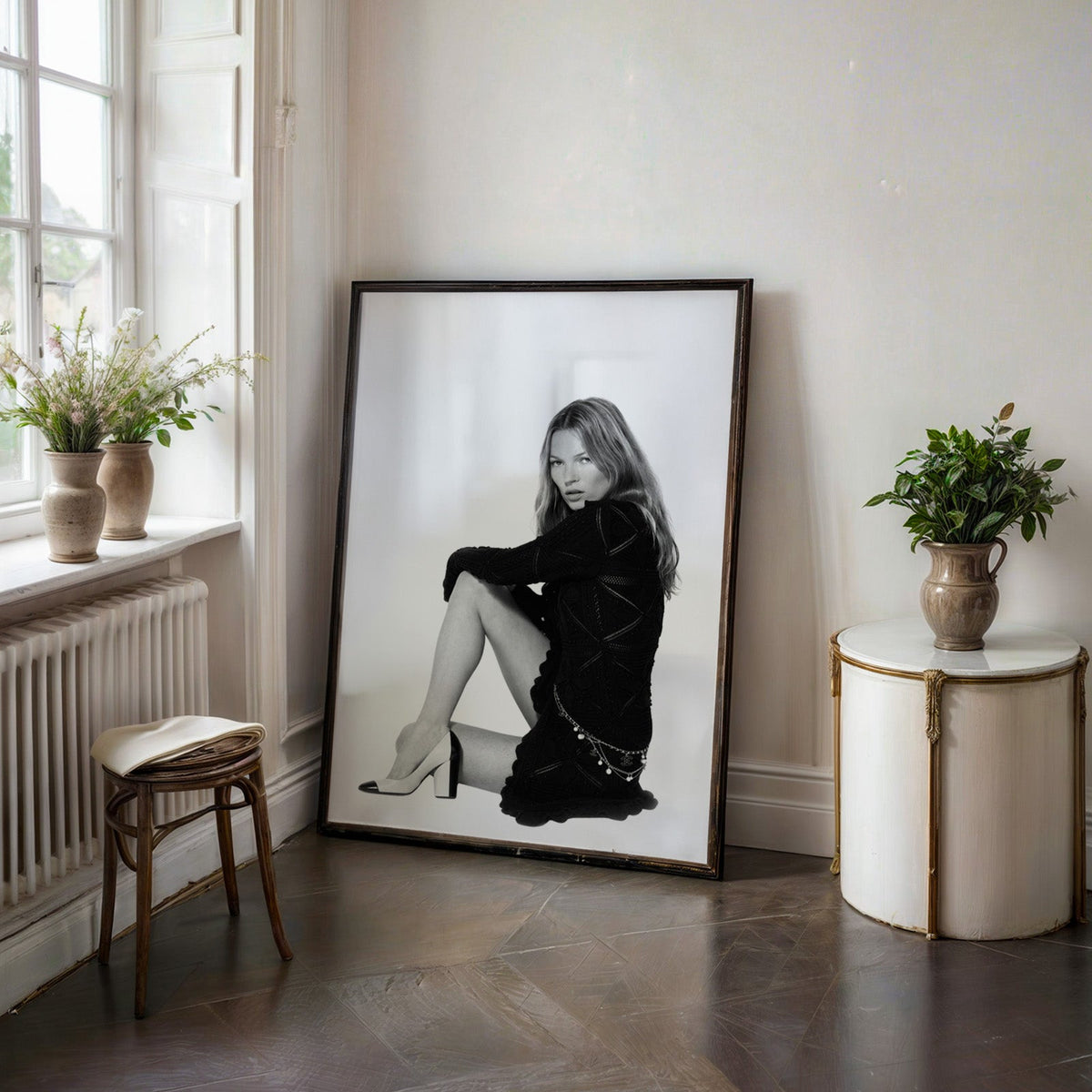 Seated woman in lace-pattern knit dress on white print in a black frame on a dark stone floor beside a white side table