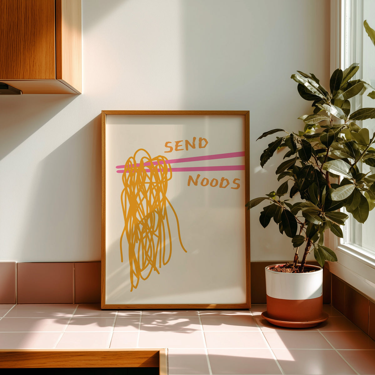 Send Noods chopsticks print in a golden oak frame leaning on a pink tiled ledge in strong afternoon sun
