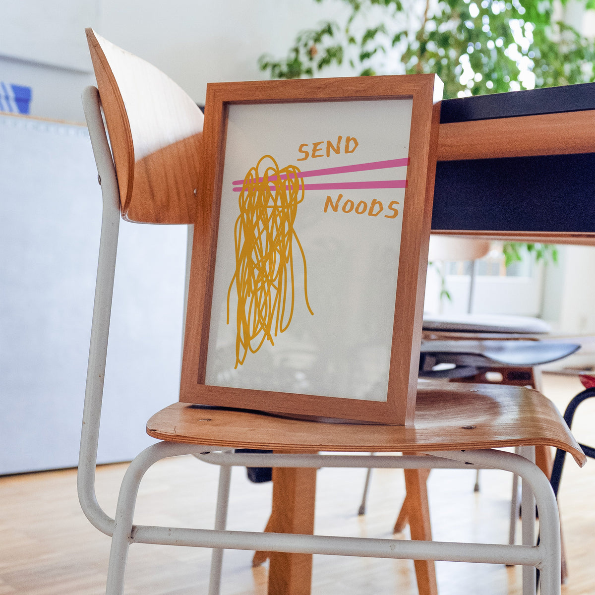Send Noods chopsticks print in a walnut frame resting on a plywood studio chair beside a dark desk