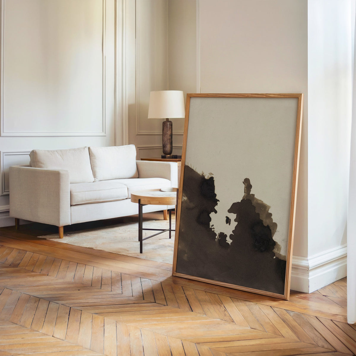 Sepia ink blot facing profiles abstract print in an oak frame leaning on a herringbone floor by a cream linen sofa
