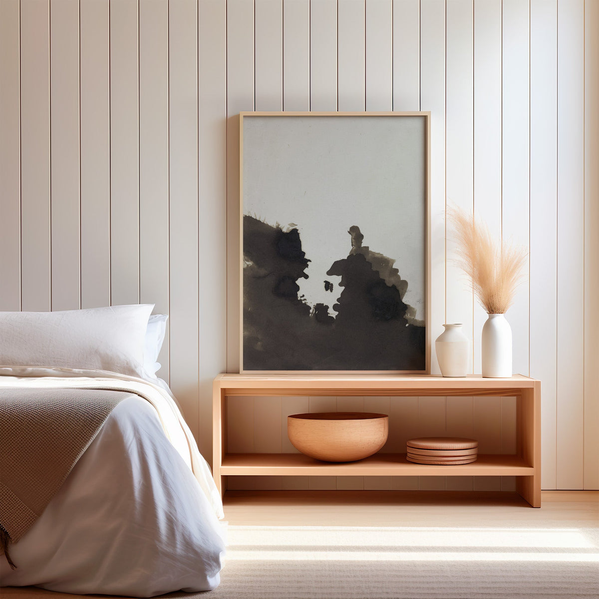 Sepia ink blot facing profiles abstract print in a light oak frame on a wood bench by a bed with pampas grass