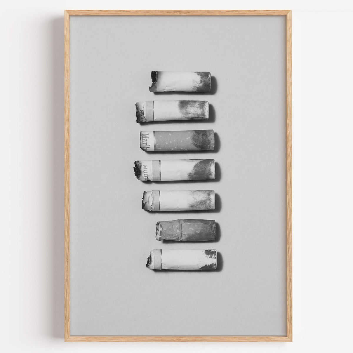 Seven cigarette ends in a column on gray print in a light oak frame on a plain white wall