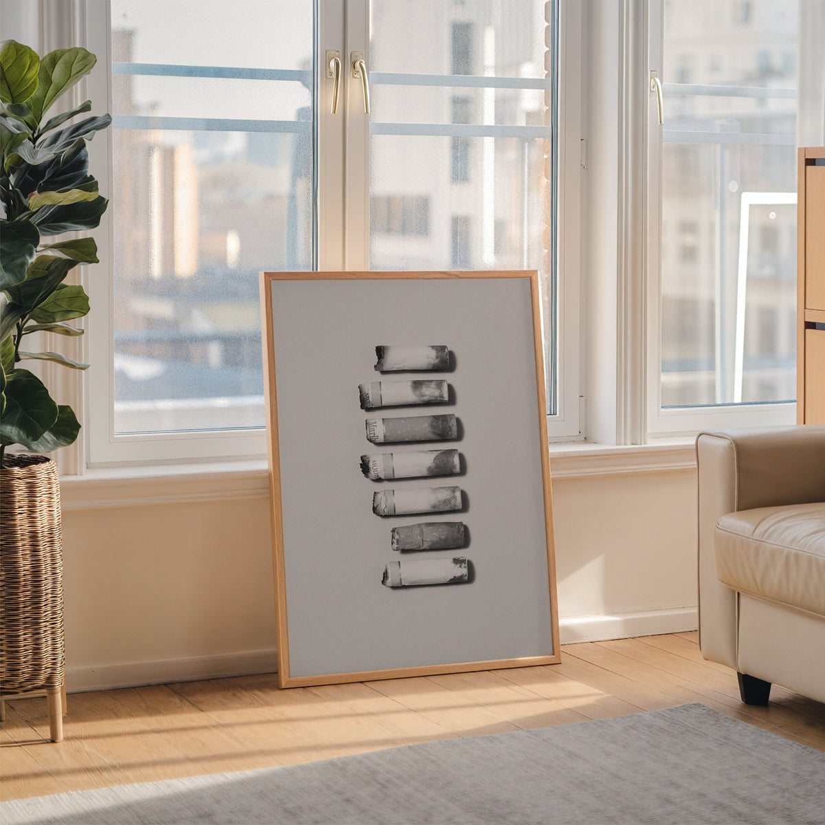 Seven cigarette ends in a column on gray print in an oak frame leaning on the floor beside a fiddle leaf fig