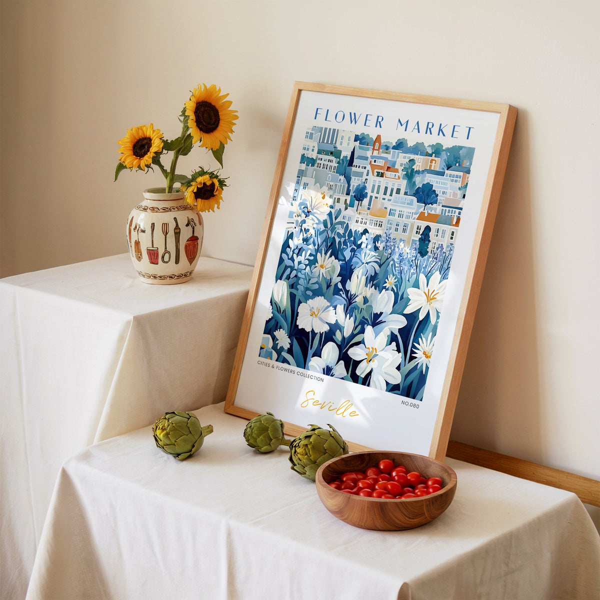 Seville flower market print in an oak frame leaning on a linen-draped table beside a vase of sunflowers