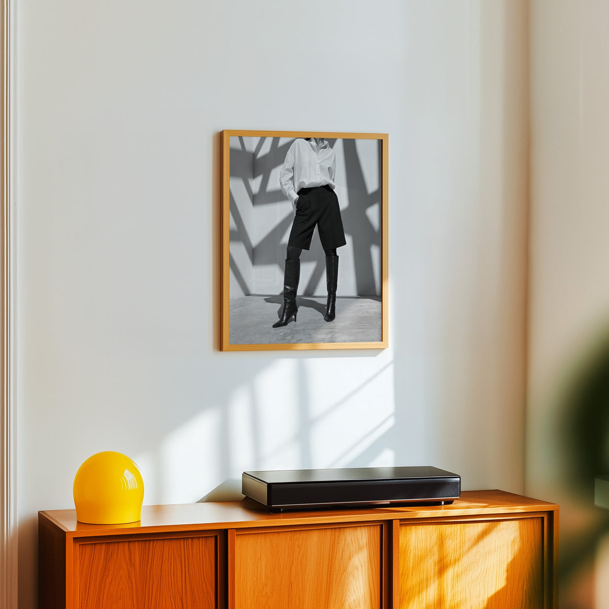 Black and white shirt and knee boots shadow wall print in an oak frame above a teak sideboard with a yellow glass lamp