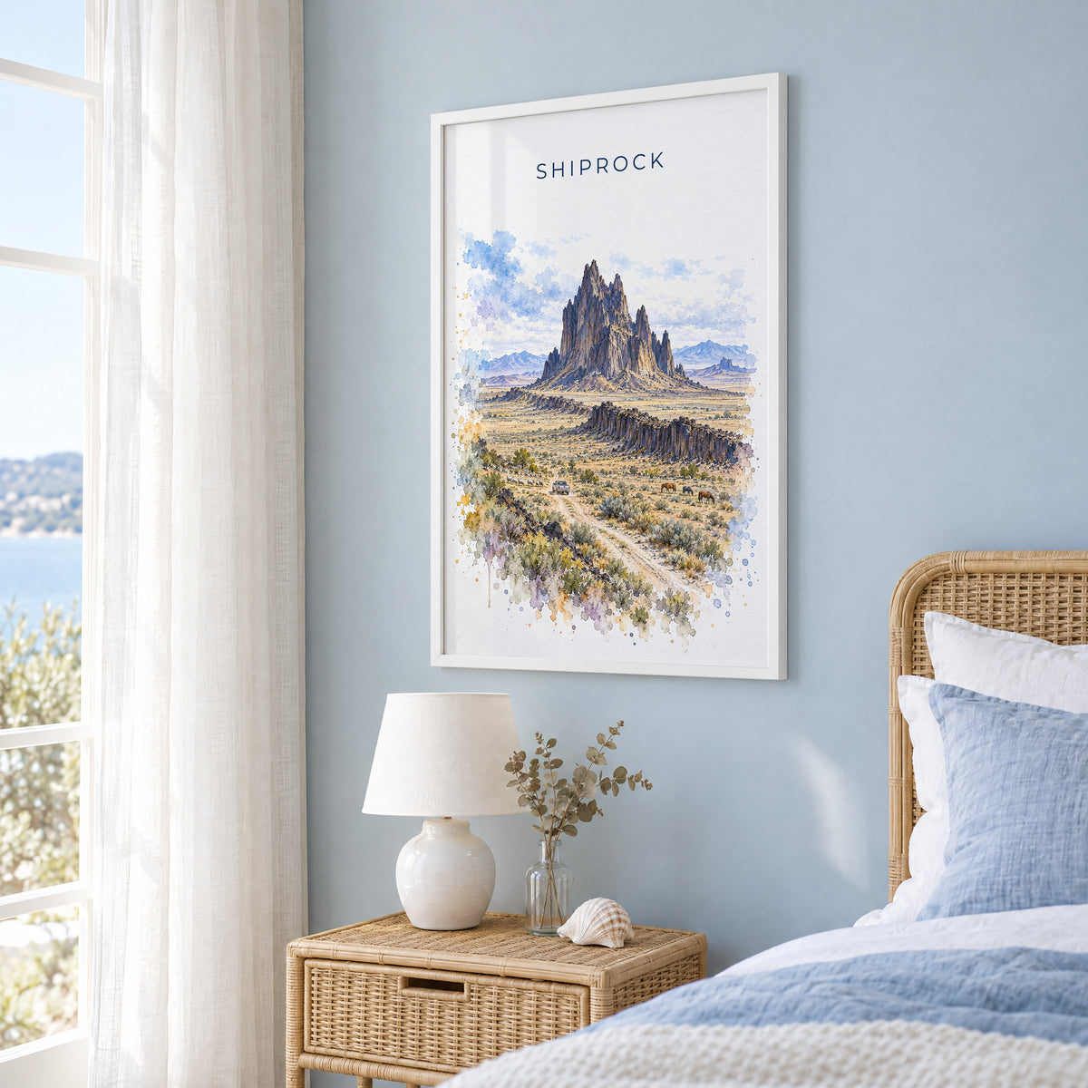 Shiprock watercolor print in a white frame on a pale blue wall beside a rattan bed in a coastal bedroom