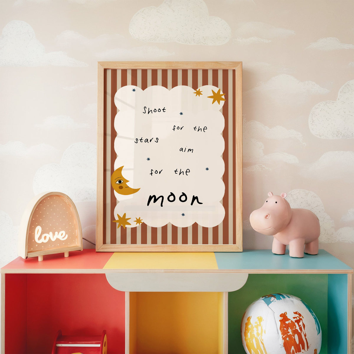 Shoot for the stars moon nursery print in an oak frame on a rainbow toy shelf against cloud wallpaper