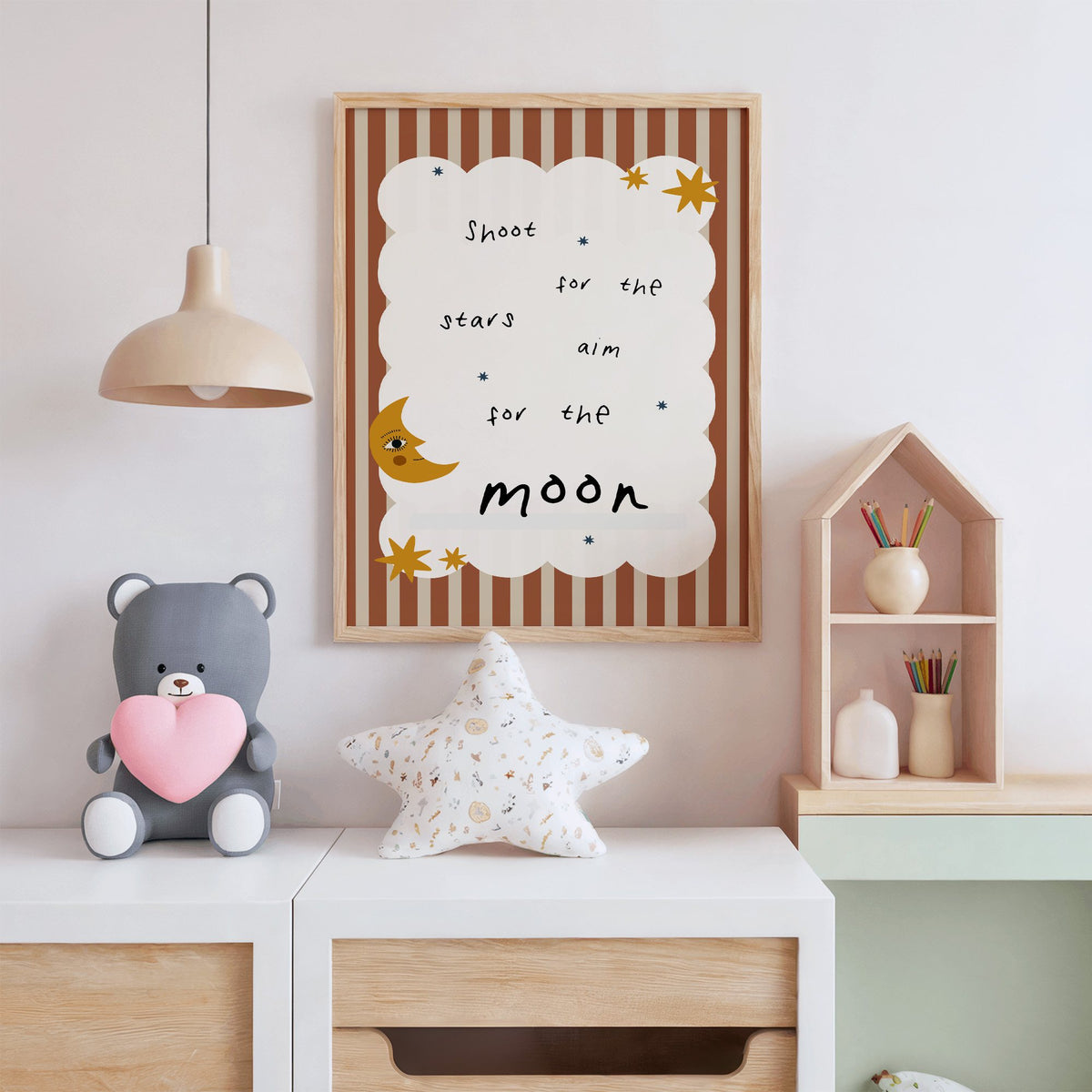 Shoot for the stars moon nursery print in an oak frame above a white dresser with a star pillow and house shelf